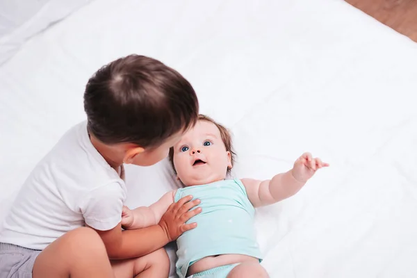 Симпатичный мальчик обнимает младшую сестру на белоснежной кровати — стоковое фото