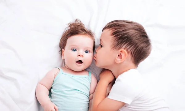 Malý krásný bratr políbí svou novorozenou sestru na tvář, zatímco leží na posteli — Stock fotografie