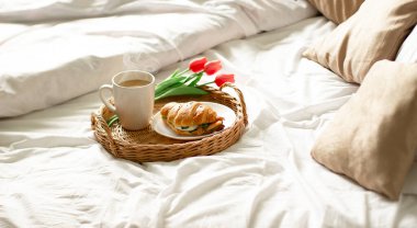 Otel odasındaki yatakta kahvaltı tepsisi. Kar beyazı bir yatakta kahvaltı, hasır tepside kruvasanla kahve. Günaydın..