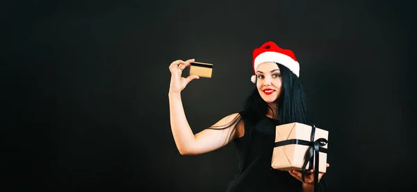 彼女の頭にクリスマスの帽子をかぶった幸せな美しい若い女性ブラック・フライデー・ホリデーの黒い背景に黒いリボンとクレジットカードを手にした贈り物。テキストの空間 — ストック写真