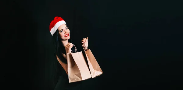 豪華なコンセプトは ブラックフライデーの休日に黒い背景にショッピングバッグを持つ彼女の頭の上にクリスマスの帽子を持つ幸せな美しい若い女性です テキストの空間 — ストック写真