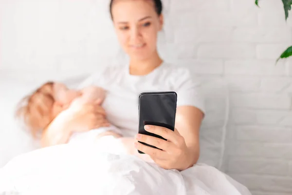 家里一个带着新生儿的年轻母亲正坐在雪白的床上 而母亲则在看着电话 — 图库照片