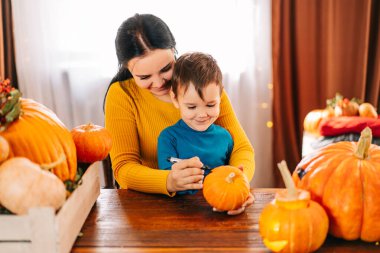 Mutlu bir aile Cadılar Bayramı için hazırlanıyor. güzel genç bir anne ve oğlu evde siyah bir kalemle bir balkabağı yüzü çiziyor.