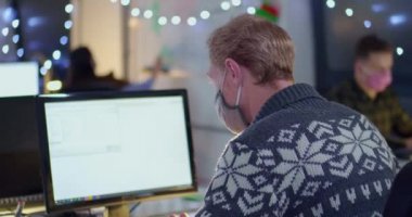 Yüz maskeli bir adam açık uzay ofisinde bilgisayarla çalışıyor.