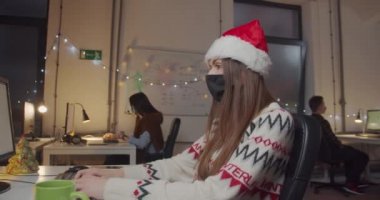 Noel Baba şapkalı ve maskeli kadın ofiste bilgisayarla çalışıyor.