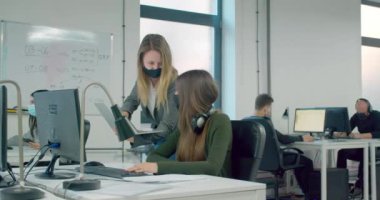 Maske takan ve bilgisayarını tutan sarışın bir iş kadını ofisteki meslektaşlarına danışıyor.