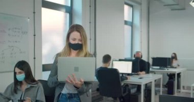 Maske takan ve bilgisayarını tutan sarışın bir iş kadını ofisteki meslektaşlarına danışıyor.
