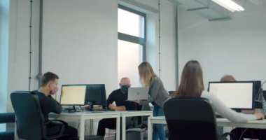 Maske takan ve bilgisayarını tutan sarışın bir iş kadını ofisteki meslektaşlarına danışıyor.