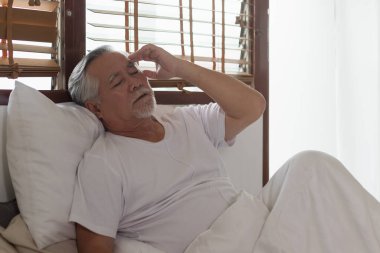 Baş ağrısı olan Asyalı yaşlı bir adam sabahları yatakta oturuyor.