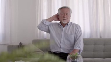 Oturma odasında kanepede oturan, başı ağrıyan Asyalı yaşlı bir adam..