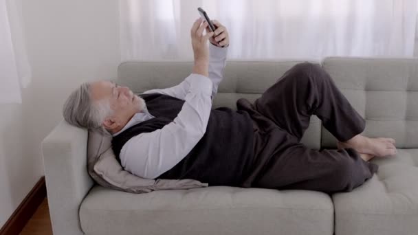 リラックスした日にソファの上でスマートフォンでアジアの高齢者の男のビデオ通話 — ストック動画