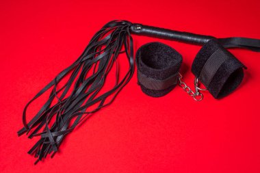 Kırmızı arka planda BDSM aksesuarları. Deri bir kırbaç ve yumuşak kelepçeler yan yana yatar. Cinsel aksesuarlar