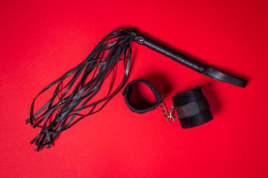 Kırmızı arka planda BDSM aksesuarları. Deri bir kırbaç ve yumuşak kelepçeler yan yana yatar. Cinsel aksesuarlar