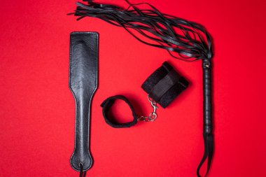 Kırmızı arka planda BDSM aksesuarları. Deri bir kürek, yastıklı kelepçeler ve kırbaç yan yana. Cinsel aksesuarlar