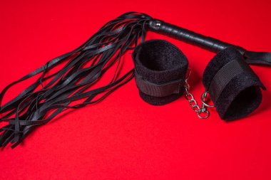 Kırmızı arka planda BDSM aksesuarları. Deri bir kırbaç ve yumuşak kelepçeler yan yana yatar. Cinsel aksesuarlar