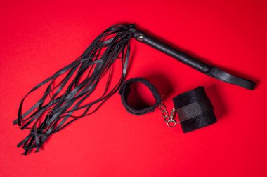 Kırmızı arka planda BDSM aksesuarları. Deri bir kırbaç ve yumuşak kelepçeler yan yana yatar. Cinsel aksesuarlar