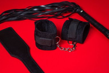 Kırmızı arka planda BDSM aksesuarları. Deri bir kürek, yastıklı kelepçeler ve kırbaç yan yana. Cinsel aksesuarlar