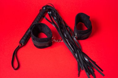 Kırmızı arka planda BDSM aksesuarları. Deri bir kırbaç ve yumuşak kelepçeler yan yana yatar. Cinsel aksesuarlar