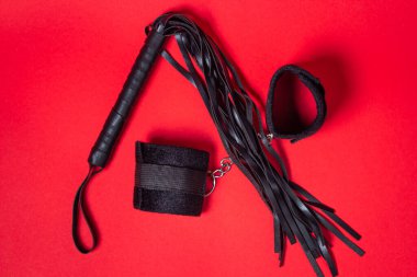 Kırmızı arka planda BDSM aksesuarları. Deri bir kırbaç ve yumuşak kelepçeler yan yana yatar. Cinsel aksesuarlar