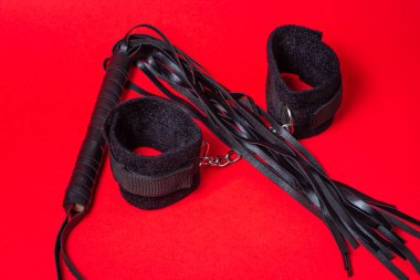 Kırmızı arka planda BDSM aksesuarları. Deri bir kırbaç ve yumuşak kelepçeler yan yana yatar. Cinsel aksesuarlar