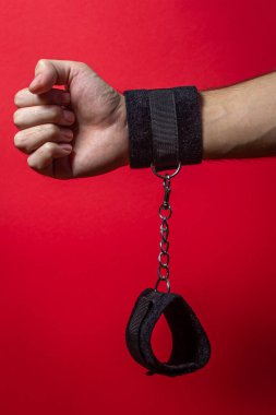 Yumuşak kelepçeler. Kırmızı arka planda elleri kelepçeli bir erkek. BDSM aksesuarları
