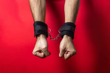 Yumuşak kelepçeler. Kırmızı arka planda elleri kelepçeli bir erkek. BDSM aksesuarları