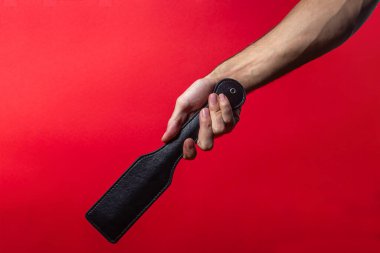 Deri kürek. Bir adamın eli kırmızı arka planda deri bir kürek tutuyor. BDSM aksesuarları