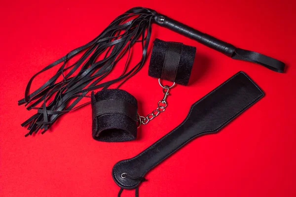 Kırmızı arka planda BDSM aksesuarları. Deri bir kürek, yastıklı kelepçeler ve kırbaç yan yana. Cinsel aksesuarlar