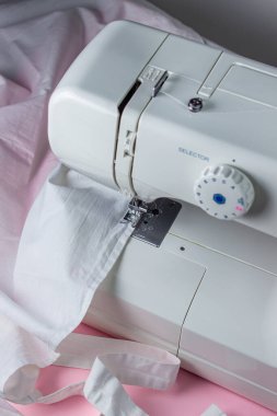 Pembe arka planda beyaz dikiş makinesi. Elektrikli dikiş makinesi. Dikiş makinesindeki beyaz kumaş. El yapımı bir alet. Dikiş işi.