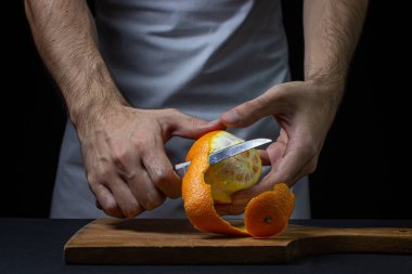 Koyu arkaplanda turuncu. Şef bir portakal soyuyor. Erkek eli siyah arka planda portakal kabuğunu kesmiş.