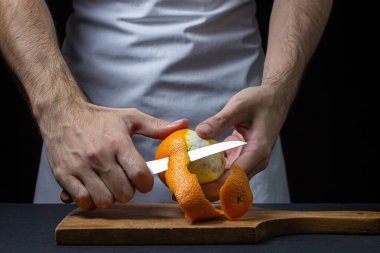 Koyu arkaplanda turuncu. Şef bir portakal soyuyor. Erkek eli siyah arka planda portakal kabuğunu kesmiş.