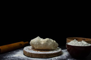 Karanlık bir geçmişi var. Siyah arka planda, tahta bir tahtanın üzerinde ev yapımı bir hamur parçası. Hamurla çalışmak için gerekli aletler hamurun etrafında yatar..