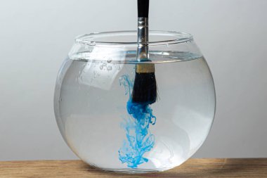 A brush with blue paint is dipped in a jar of water on a white background. The paint spreads in the water. clipart