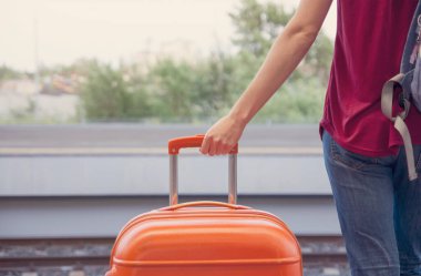Genç bir kadın, tren istasyonunun peronunda büyük turuncu bir bavul tutuyor.