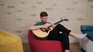 Genç adam gitar çalıyor. Oturma odasında kırmızı bir çantanın üzerinde oturuyor..