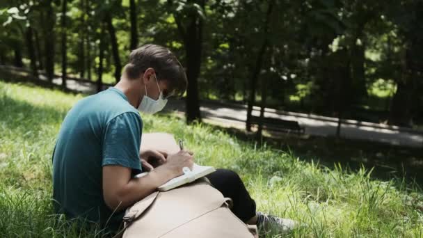 Homem com uma máscara médica na cara sentado no parque e escrever novas ideias canção. — Vídeo de Stock