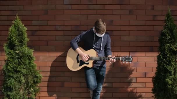 Młody człowiek w leczniczej masce gra na gitarze na zewnątrz.. — Wideo stockowe