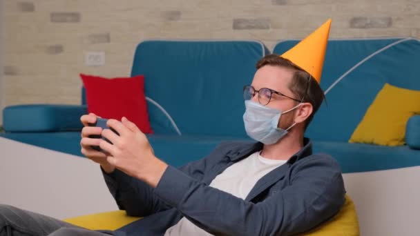 Een jongeman met een verjaardagshoed en een medisch masker op zijn gezicht praat online met zijn vrienden. — Stockvideo