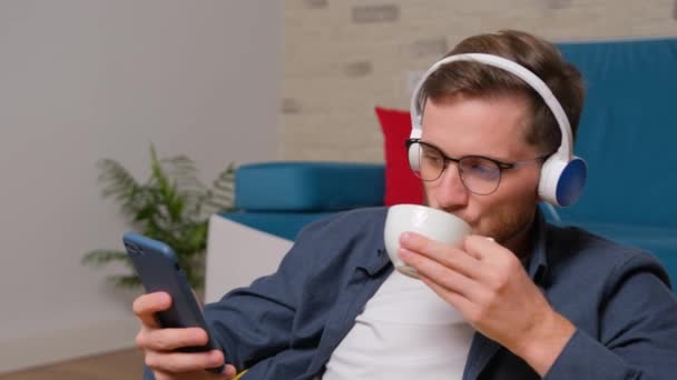 Joven escuchando música en los auriculares y bebiendo una taza de café. — Vídeo de stock