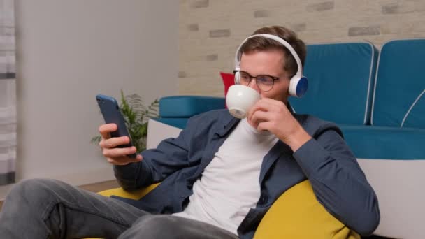 Joven escuchando música en los auriculares y bebiendo una taza de café. — Vídeo de stock