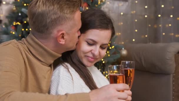 Pareja enamorada sentada y abrazada cerca del árbol de Navidad mientras bebe champán. — Vídeos de Stock