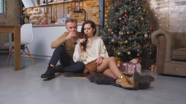 Pareja joven bebiendo champán mientras se sienta y se abraza cerca del árbol de Navidad. — Vídeos de Stock