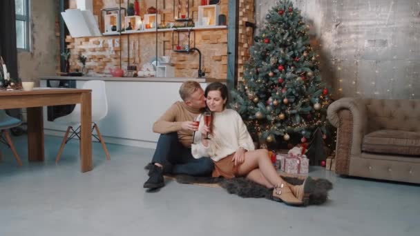 Casal apaixonado sentado e abraçando perto da árvore de Natal enquanto bebe champanhe. — Vídeo de Stock