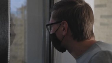 Koronavirüs karantinası sırasında maskeli adam pencereden dışarı bakıyor..