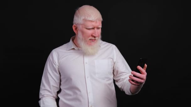Homem engraçado com albinismo usando telefone e falando on-line no fundo preto. — Vídeo de Stock
