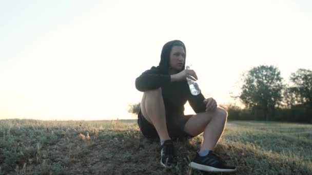 Sportieve man neemt een pauze van de sport, zitten op gras en drink een fles water. — Stockvideo