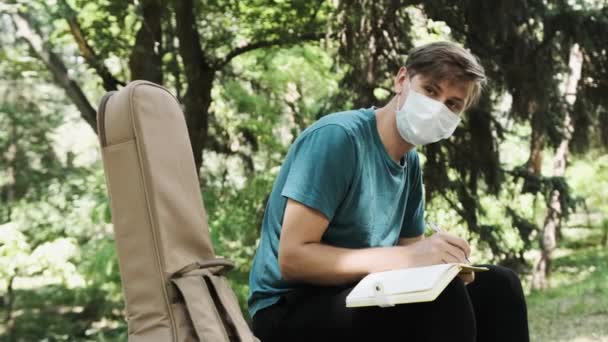 El joven está sentado en un banco en el parque y escribe algunas ideas de canciones en un cuaderno.. — Vídeos de Stock