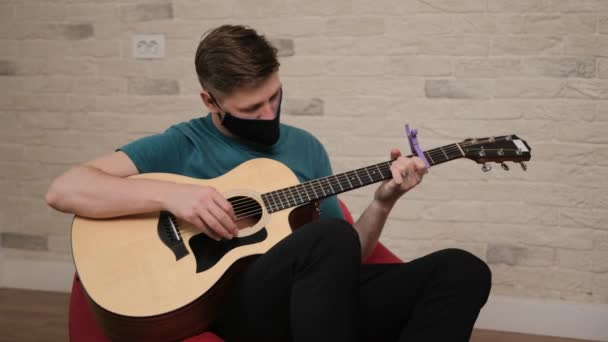 Blanke man met een zwart medisch masker op het gezicht speelt op de gitaar — Stockvideo