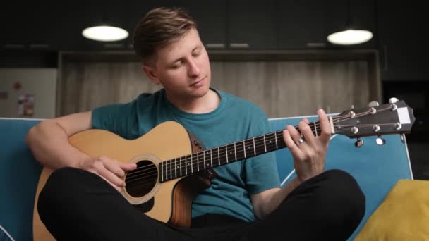 Молодой человек, сидящий на синем диване и играющий на гитаре, наслаждается жизнью. — стоковое видео