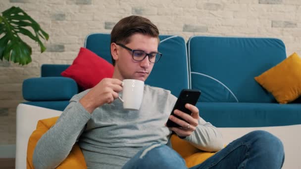 El hombre escribe un mensaje usando un teléfono móvil y bebiendo una taza de té. — Vídeos de Stock